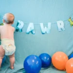 baby in white diaper standing beside blue and red balloons