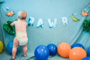 baby in white diaper standing beside blue and red balloons