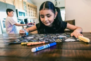 a little girl sitting at a table with crayons and markers