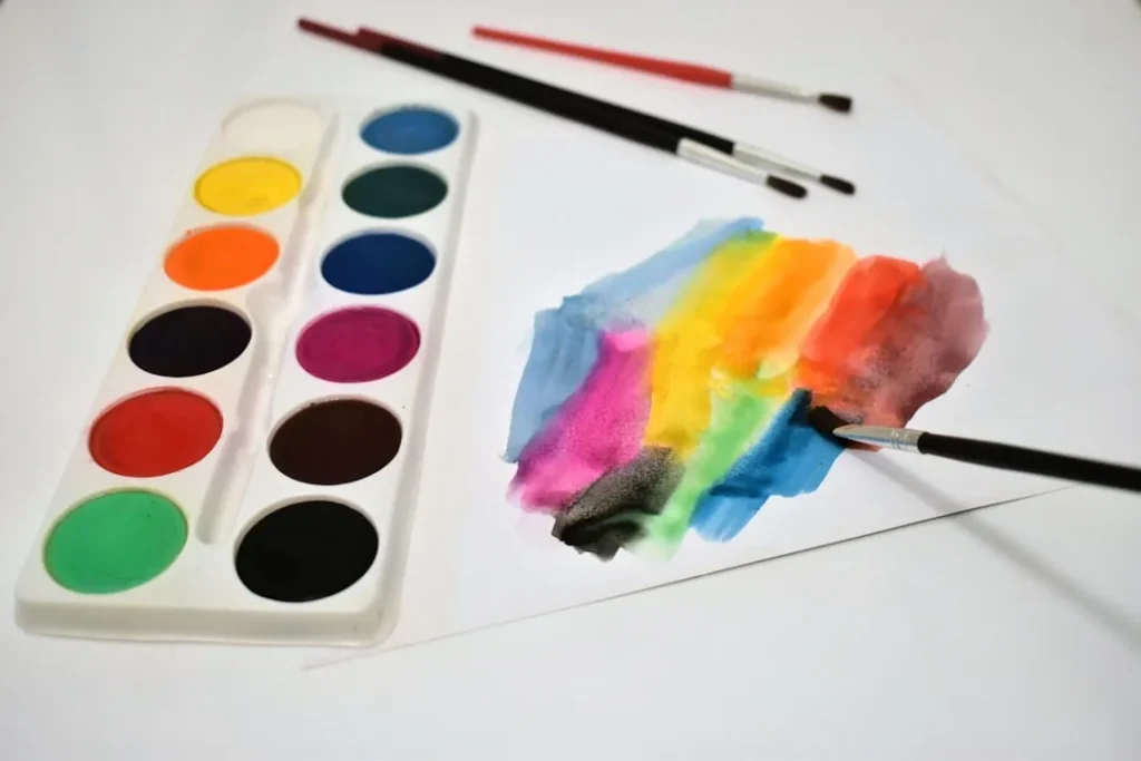 Watercolor paint set, brushes, and colorful strokes on white paper.